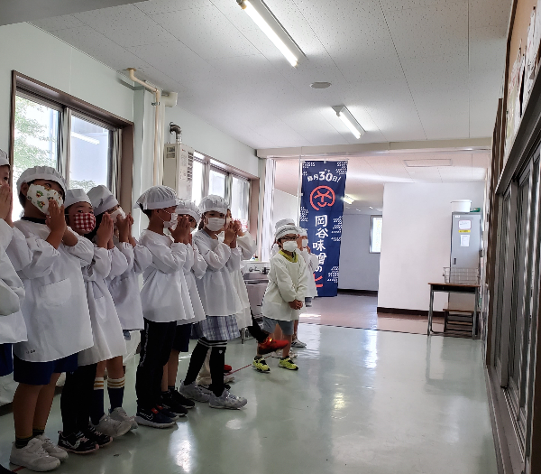 給食室前であいさつをする給食当番の児童と岡谷味噌のヒと書かれた桃太郎旗の写真