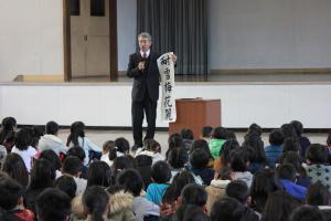 体育館で座っている子どもたちの前で「耐雪梅花麗」と書かれた紙を持ち話をする男性の写真