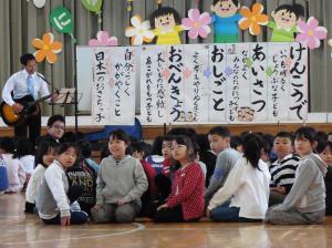 体育館ステージ前でギターを弾いている男性を座ってみている子どもたちの写真
