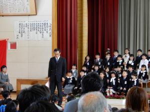 体育館のステージに子どもたちが座って並んでいる前で1人の男性が立っている写真