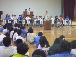 体育館で演台前にたってスピーチしているのを座ってみている子どもたちの写真