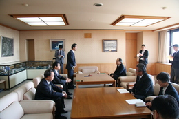 会議室内で記者に見守られながらスーツ姿の数名が調印式に臨んでいる写真