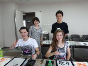 笑顔で筆を執る外国人参加者の男女二人とその後ろに立つ日本人の男女二人の写真