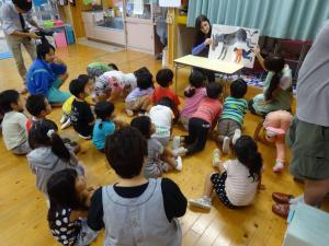 あやめ保育園にて、外国人女性の参加者が座っている園児たちに英語の絵本を読み聞かせている写真