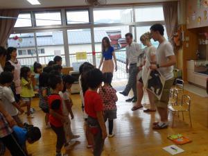 あやめ保育園にて、外国人女性の参加者が立っている園児たちに英語の絵本を読み聞かせている写真