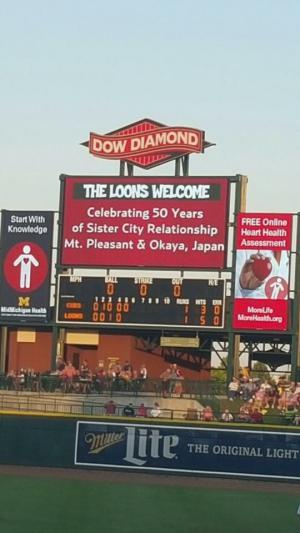 野球場のスコアボードと、「ようこそ、ルーンズへ。祝、マウント・プレザント市と岡谷市の姉妹都市友好50周年」と書かれた看板の写真