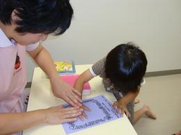 テーブルの上で女性の手助けをされながら紙に手形を取っている子どもの写真