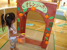 小さな女の子がオレンジの木と書かれたアーチからオレンジを取ってバスケットに入れている写真