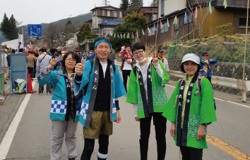道路上で男女4人が水色と緑のはっぴを着て記念撮影している写真