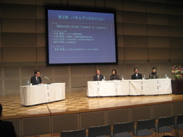 スクリーンのあるステージ上の机に座り話す男性と四人のパネリストの写真