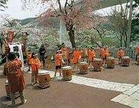 桜がきれいに咲いている広場でオレンジ色の法被を着た方たちが腰の高さの太鼓をたたいている様子の成田公園の写真