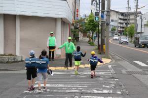 黄緑色のジャンパーを着て、黄色い旗をもった見守り隊の方が横断歩道を渡る児童を誘導する様子の写真