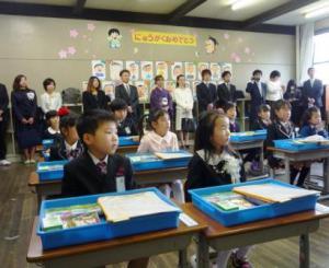 入学式で教室の後ろで父兄が並んで立っている前で新一年生たちが話を聞いている写真