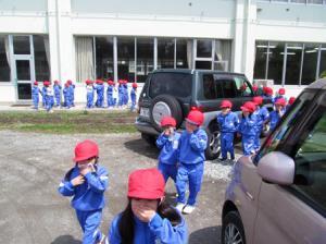 運動服を着た子どもたちが口を手で押さえながら車の間を抜けて校庭へ向かって歩いている写真