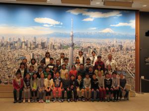 修学旅行にて東京スカイツリーを中心とした東京の街並みが描かれた絵の前で6年生の子どもたちと先生たちが並んでいる集合写真