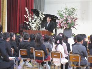 ピアノと花が置かれている舞台の上に立つ男性と、椅子に座っている一年生たちの写真
