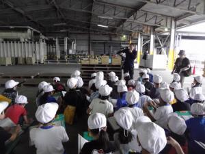 白い帽子をかぶった生徒たちが工場見学で男性から説明を座って聞いている写真