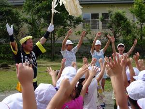 御柱休めの行事で中心役をしている一人の男性とたくさんの子どもたちが両手を上に挙げている写真