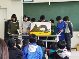 複数人の生徒たちが黒板の前で紙芝居を読んでいる写真