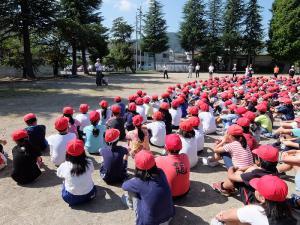 校庭で赤い帽子をかぶった子どもたちが体育座りをしている写真