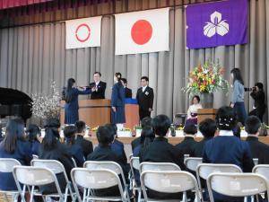 校章と日本の旗が掲げられたステージで校長先生が卒業生に卒業証書を授与している写真