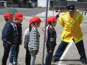 赤い運動帽子を被ったどもたちが警察の方に道の渡り方を教わっている写真