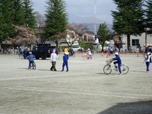 校庭の模擬道路で子どもたちが交通安全協会の方に自転車の正しい乗り方を教わっている写真