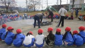 校庭で円を組んで焼きいもを焼く様子を見ている生徒たちの写真