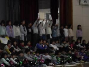 ステージの上で座っている子どもたちと立って紙を掲げている子どもたちの写真