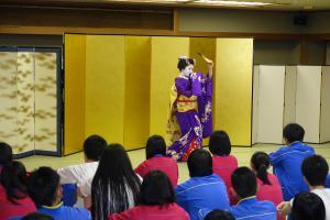 金屏風の前で和服を着て日本舞踊を踊っている人を生徒たちが座って観ている写真