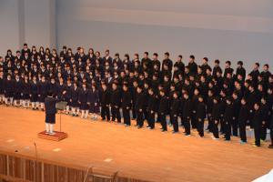ステージ上の指揮者の前で大勢の学生が左右に男女で分かれて合唱をしている写真