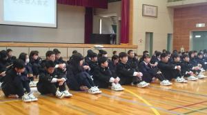 学友会入会式で新入生たちが体育座りをして真剣な表情で話を聞いている写真