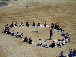 青空給食で素晴らしい天気の中、グラウンドで生徒たちが先生を囲んで座って給食を食べている写真